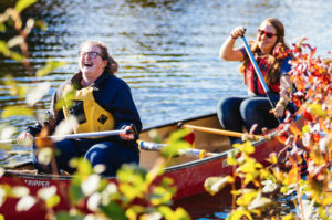 Two women laughing while paddling a canoe on a sunny river; authentic outdoor recreation and adventure lifestyle photography in Thunder Bay.