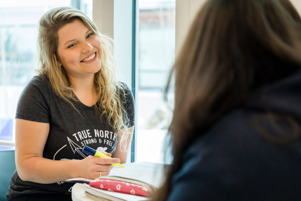 A candid shot of a Lakehead University student smiling while studying with a peer; she is wearing a "True North Strong & Free" t-shirt, capturing a friendly and collaborative academic atmosphere in Thunder Bay.