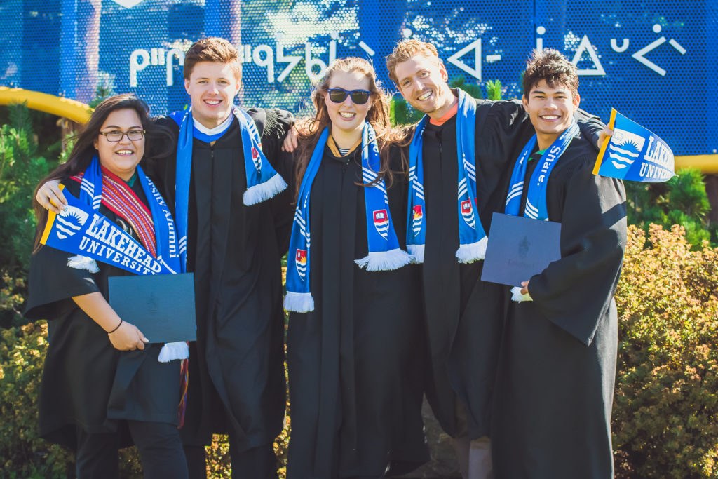 Portrait of five smiling Lakehead University graduates in black gowns and blue alumni scarves holding diplomas; professional graduation photography in Thunder Bay.