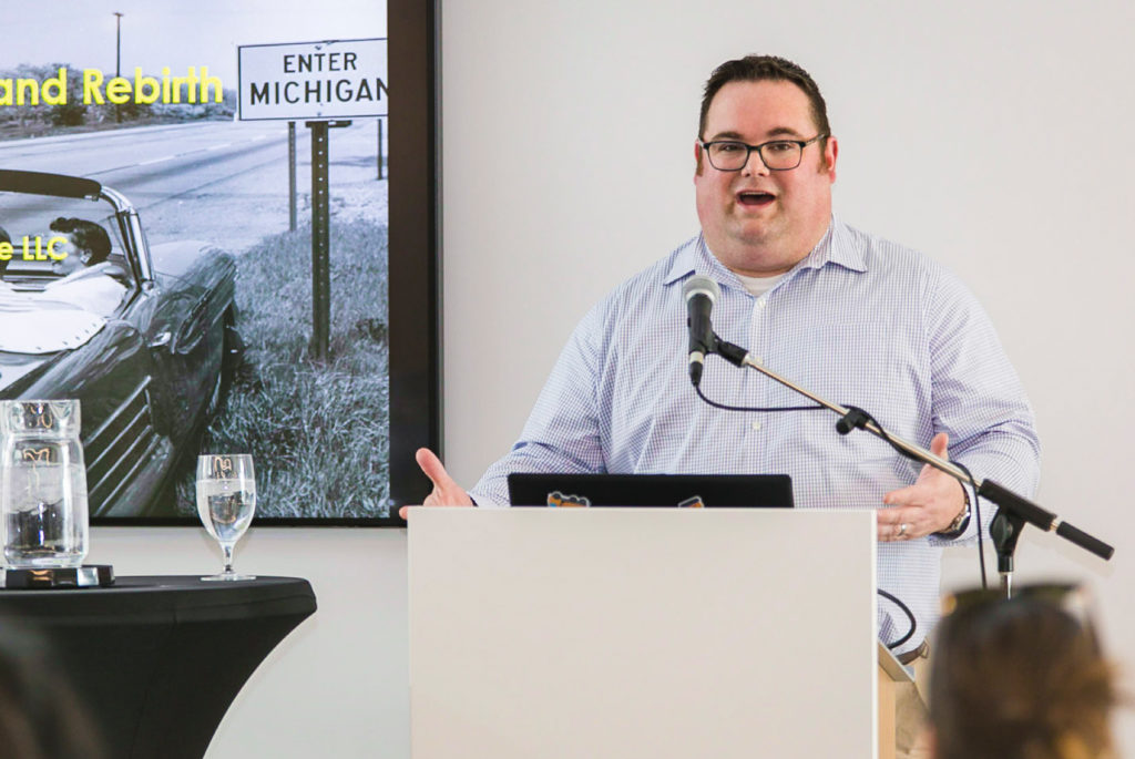 Professional speaker at a podium giving a presentation; business event photography featuring a digital screen with "Enter Michigan" slide in Thunder Bay.