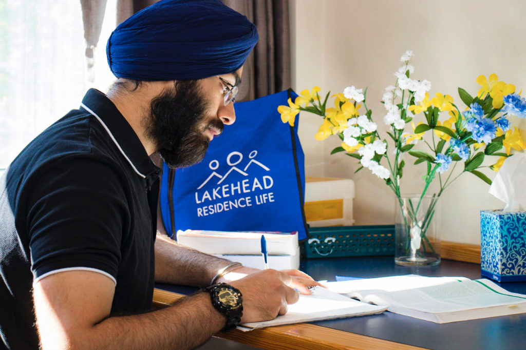 Lakehead University student with a blue turban studying at a desk in a residence room; candid campus lifestyle photography featuring "Residence Life" branding in Thunder Bay.