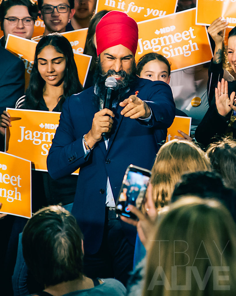A dynamic shot of Canadian politician Jagmeet Singh speaking at a rally in Thunder Bay; he is pointing toward the crowd while surrounded by supporters holding orange "Jagmeet Singh" campaign signs.