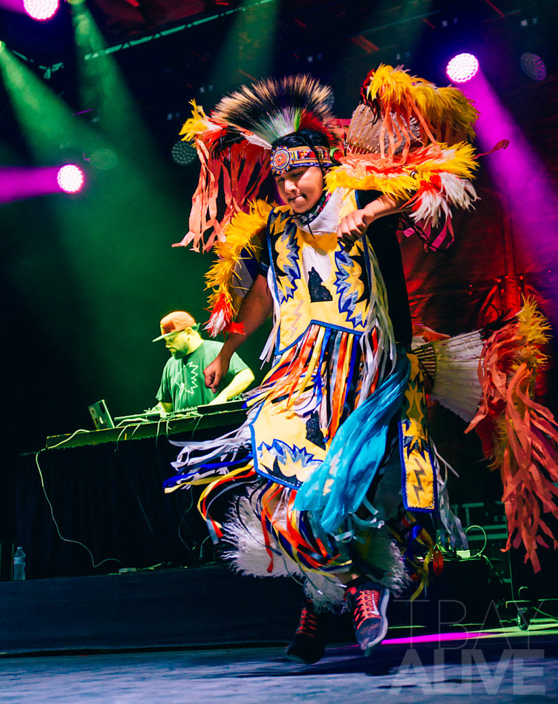 An energetic, high-contrast photograph of an Indigenous dancer in vibrant traditional regalia performing on stage at the Wake the Giant Music Festival in Thunder Bay. The shot captures fluid movement and cultural storytelling under dramatic pink and green stage lighting, with a DJ visible in the background.