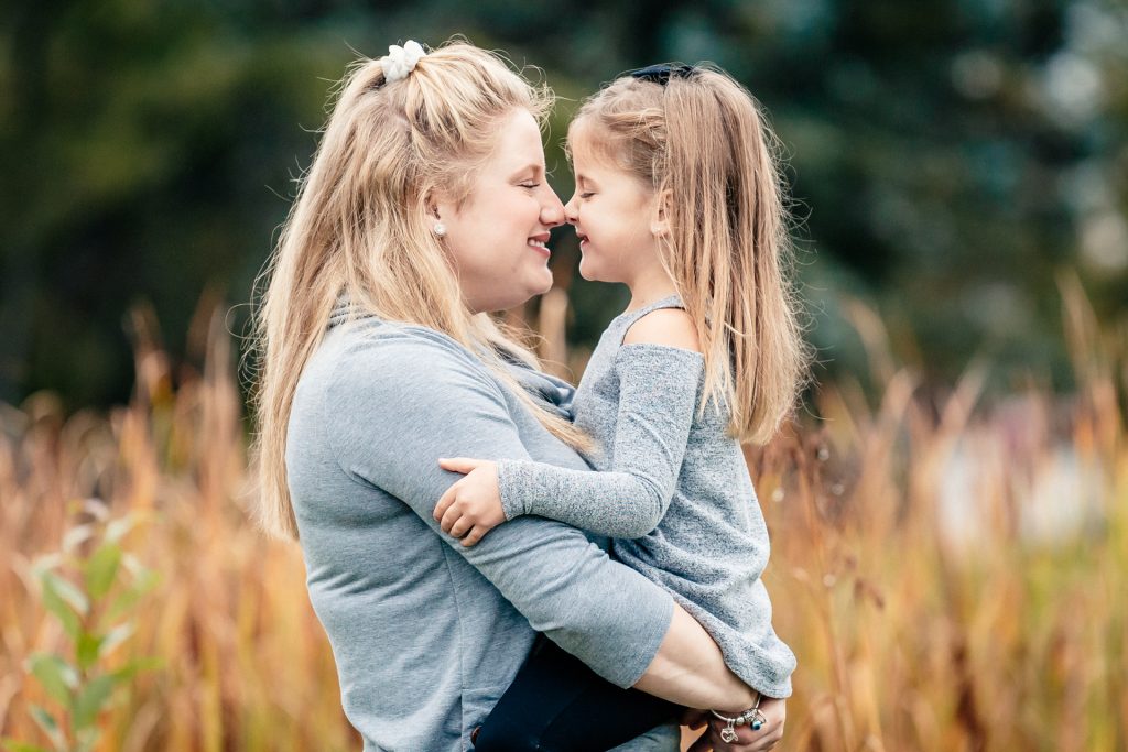 A portrait of a mother holding her young daughter, capturing a tender moment of connection as they touch noses and smile. The high-clarity shot features natural skin tones and a soft depth of field against a vibrant autumnal background in Thunder Bay, reflecting TbayAlive’s focus on authentic storytelling and genuine family interactions.