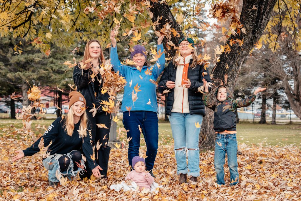 A family portrait capturing a multi-generational group and a young child playing in falling leaves at Friendship Gardens in Thunder Bay. The high-clarity shot features natural skin tones and a vibrant autumnal background, reflecting TbayAlive’s focus on authentic storytelling and the joyful, real-life moments of the local community.