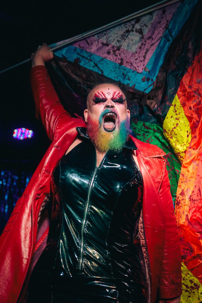 An energetic and expressive drag performance photograph capturing a performer in a black leather outfit and red jacket with a vibrant, rainbow-colored beard; the high-contrast composition highlights authentic storytelling and genuine community connection, reflecting TbayAlive’s commitment to supporting Thunder Bay’s LGBTQ+ performers and local arts scene.