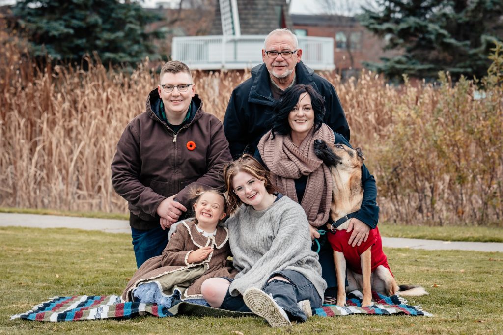 A family portrait capturing a multi-generational group and their dog sharing a genuine moment of connection at Friendship Gardens in Thunder Bay. The high-clarity shot features soft, natural skin tones and a vibrant autumnal background, reflecting TbayAlive’s focus on authentic storytelling and the real emotions of the local community.