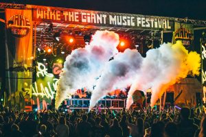 An energetic, atmospheric wide shot of the Wake the Giant Music Festival stage in Thunder Bay, featuring dramatic plumes of white smoke, vibrant stage lighting, and a dense, engaged crowd; the high-contrast composition captures the immersive community energy of the event, reflecting TbayAlive’s signature storytelling and decade-plus experience in the local live music scene.