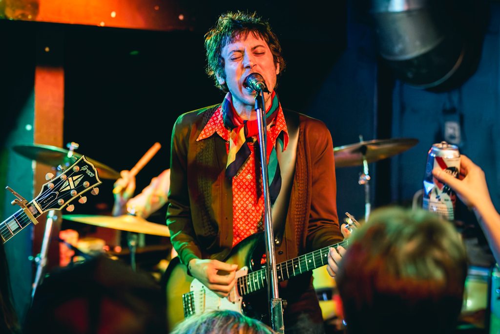 An energetic, high-contrast concert photograph of a male musician performing with intense emotion in a local Thunder Bay venue. The shot captures the raw energy of the performance through atmospheric lighting and cinematic color tones, reflecting TbayAlive’s decade-plus experience in live music photography and commitment to authentic storytelling