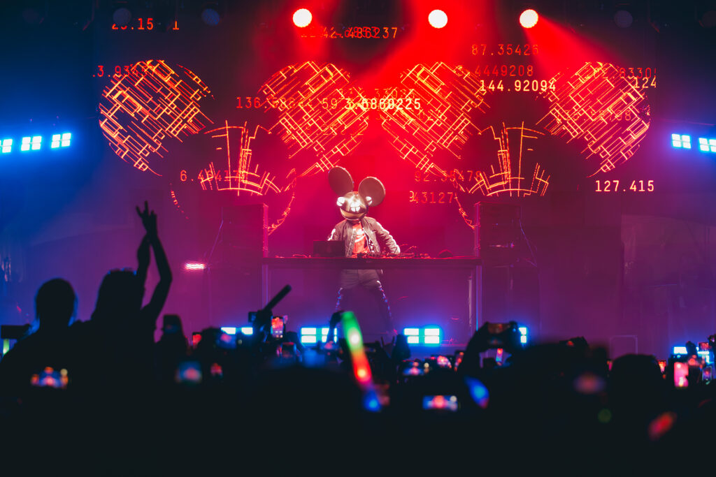 An energetic, high-contrast concert photograph of deadmau5 performing in a mouse helmet behind a DJ booth, featuring vibrant red stage lighting and dramatic digital displays. The shot captures the immersive atmosphere of the crowd in silhouette with glowing phone screens, reflecting TbayAlive’s signature cinematic storytelling and expert use of light.