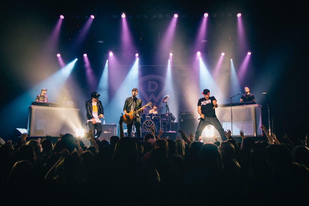 An energetic and atmospheric wide shot of a live concert performance featuring a full band on stage under vibrant purple and white spotlights; the high-contrast composition captures the immersive crowd interaction and intense stage energy, reflecting TbayAlive’s signature cinematic storytelling and decade-plus experience in Thunder Bay’s music scene