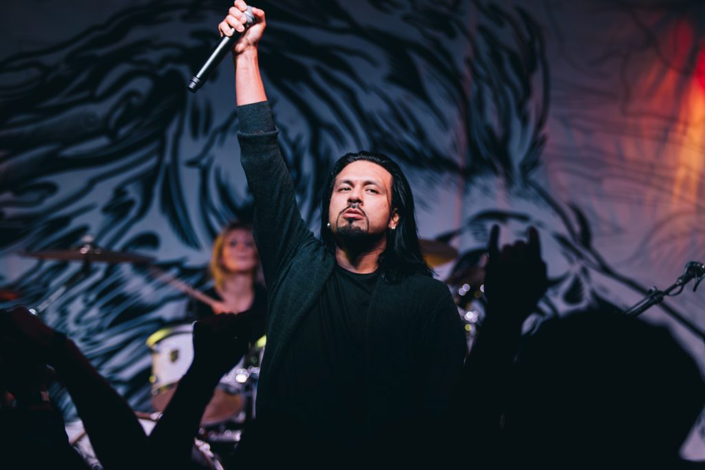 An energetic and atmospheric concert photograph of a male vocalist performing with a raised arm under dramatic stage lighting; the shot captures raw stage presence and authentic storytelling, reflecting TbayAlive’s signature cinematic style and decade-plus experience in Thunder Bay’s live music scene.