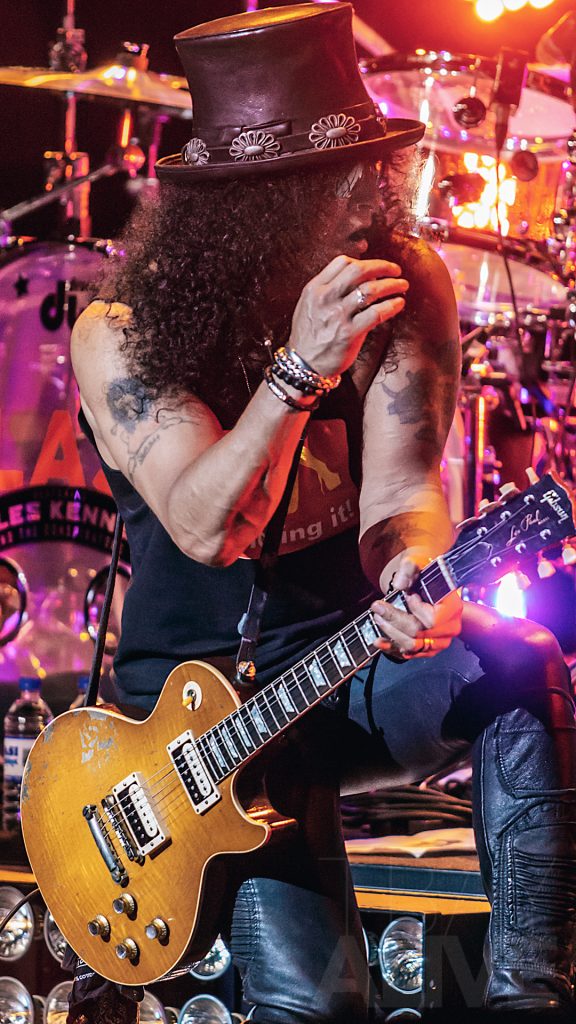 A high-contrast, energetic concert photograph of Slash performing with a Gibson Les Paul guitar while wearing his iconic top hat; the atmospheric shot captures vibrant stage lighting and raw performance energy, reflecting TbayAlive’s signature cinematic storytelling and over 10 years of experience in Thunder Bay’s live music scene.