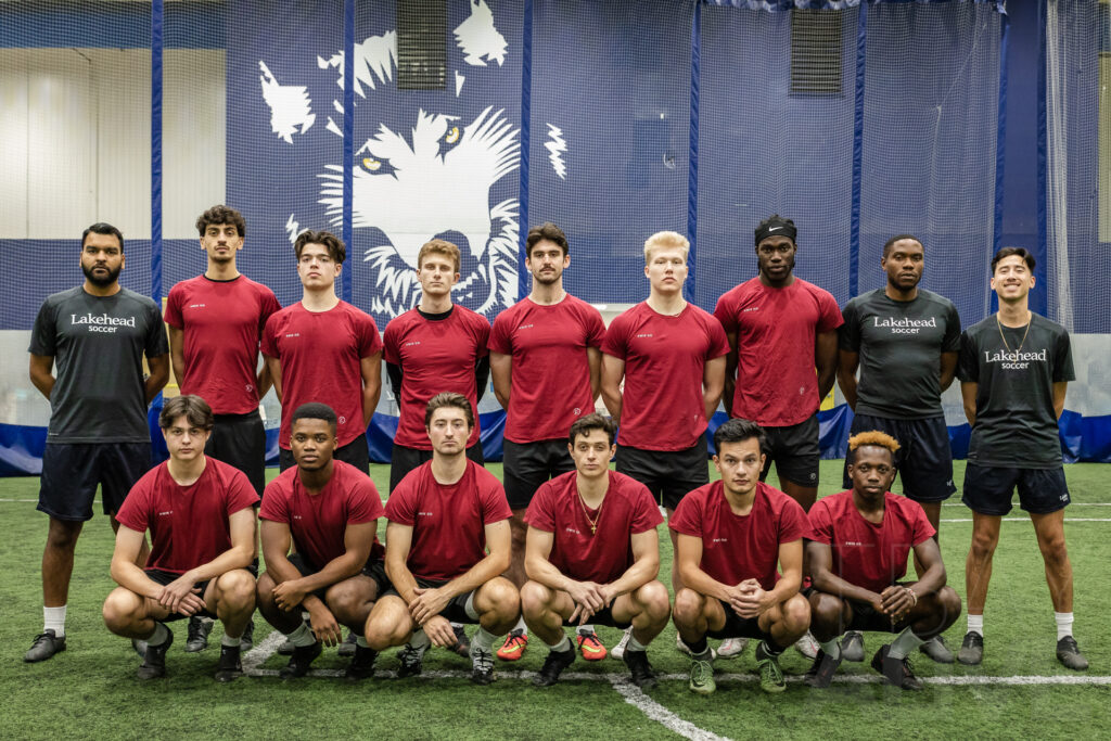 Lakehead Thunderwolves men's soccer team posed for a group photo indoors in front of a large wolf mascot banner, Thunder Bay