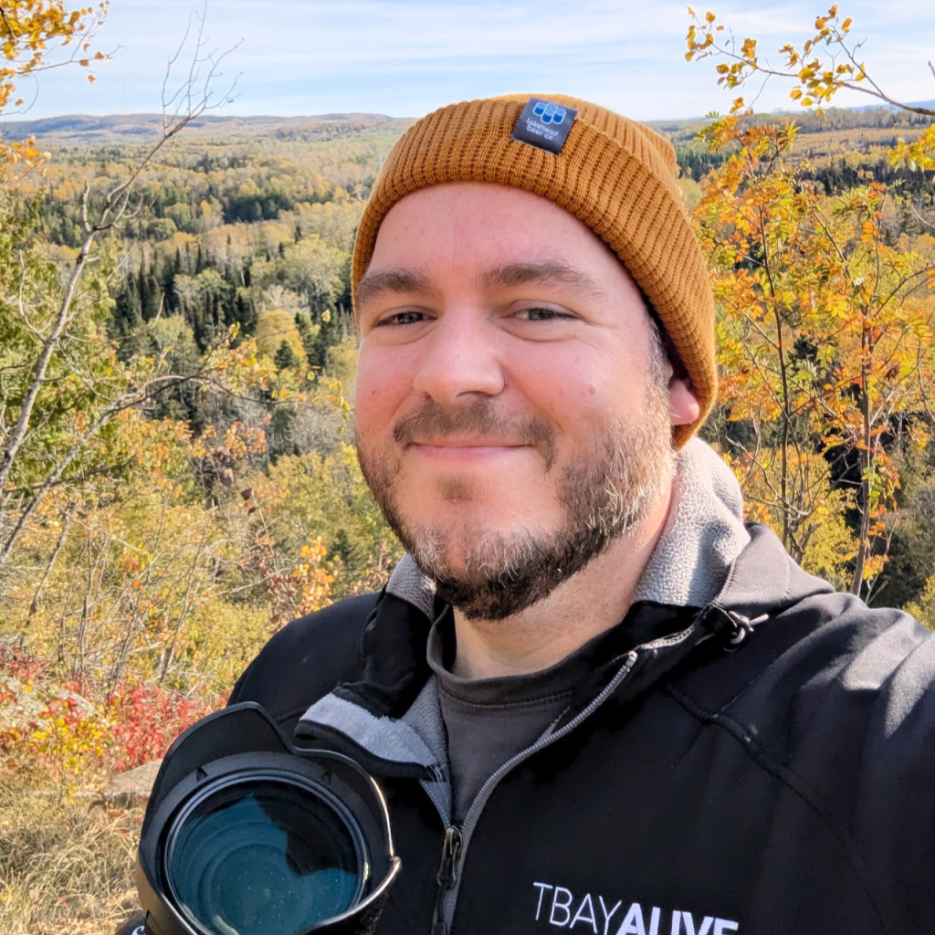 A high-clarity, approachable outdoor portrait of Cody Angus, owner of TbayAlive, wearing a branded jacket and an orange beanie while holding a camera. The background showcases the vibrant, deep hues of a Thunder Bay boreal forest in autumn, reflecting the brand’s signature cinematic color palette and passion for local Northern Ontario landscapes.