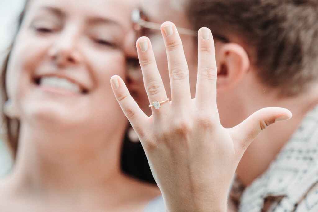 Close-up engagement portrait of a smiling woman showing her engagement ring, with her partner in the background