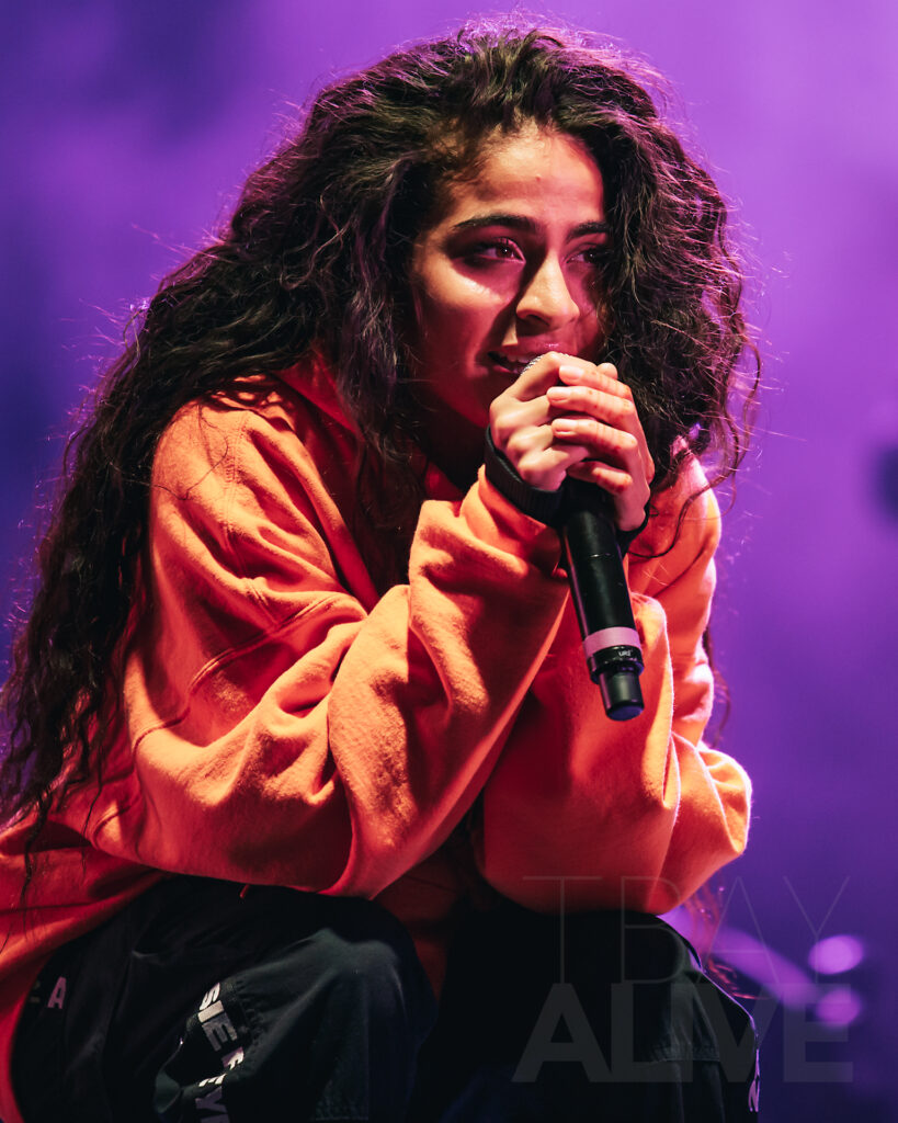 Female concert performer singing intensely into a microphone under purple stage lighting, wearing an orange hoodie, Thunder Bay concert photography