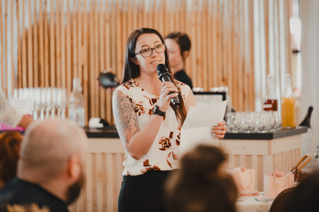 Woman speaking at a microphone at a high tea event in Thunder Bay, holding notes in a modern venue with wooden decor